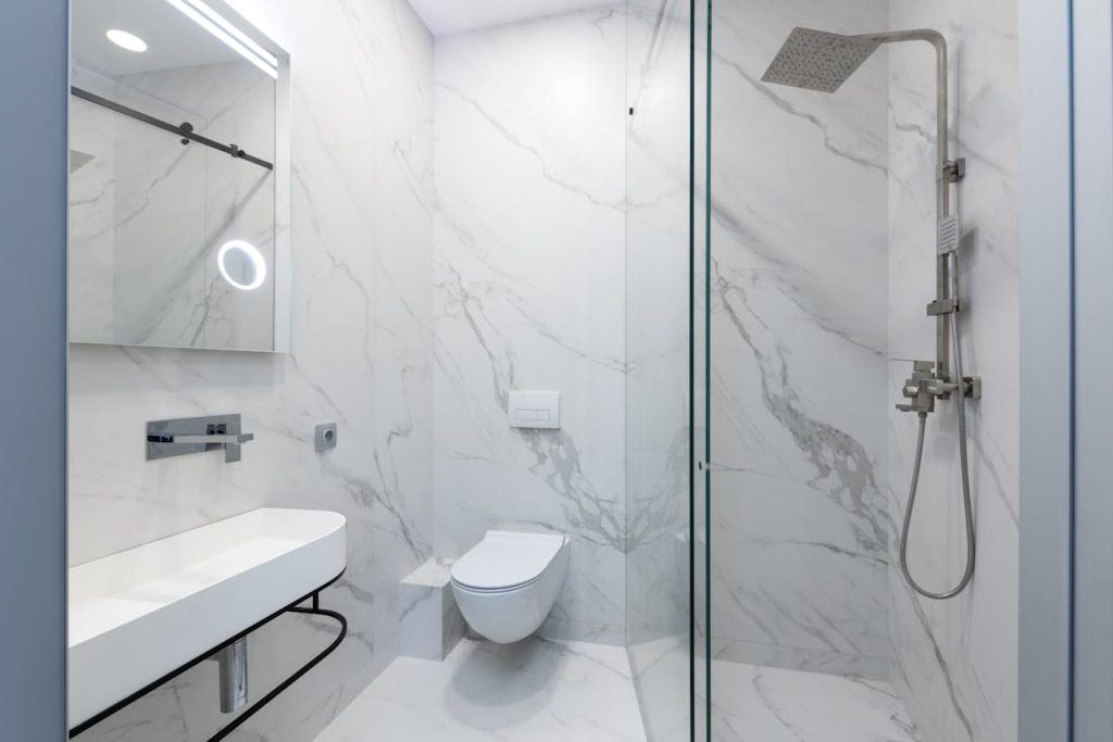 a-toilet-with-stainless-steel-shower-6444256 Elegant white marble bathroom featuring a sleek shower, toilet, and sink.